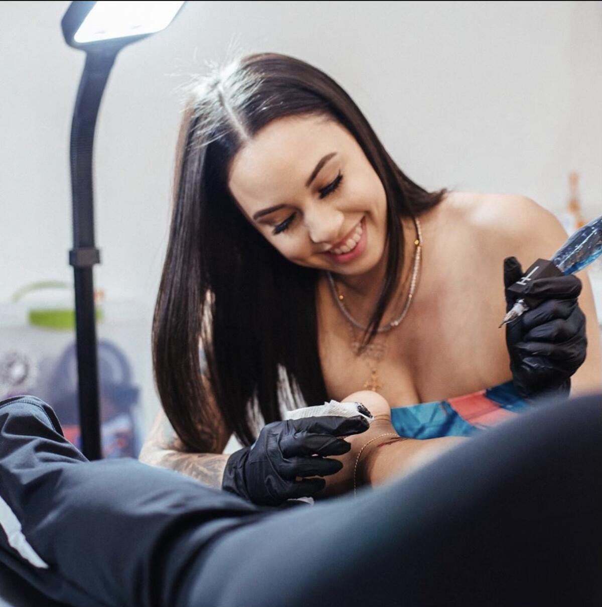 A smiling female tattoo artist with long dark hair inks up a customer 