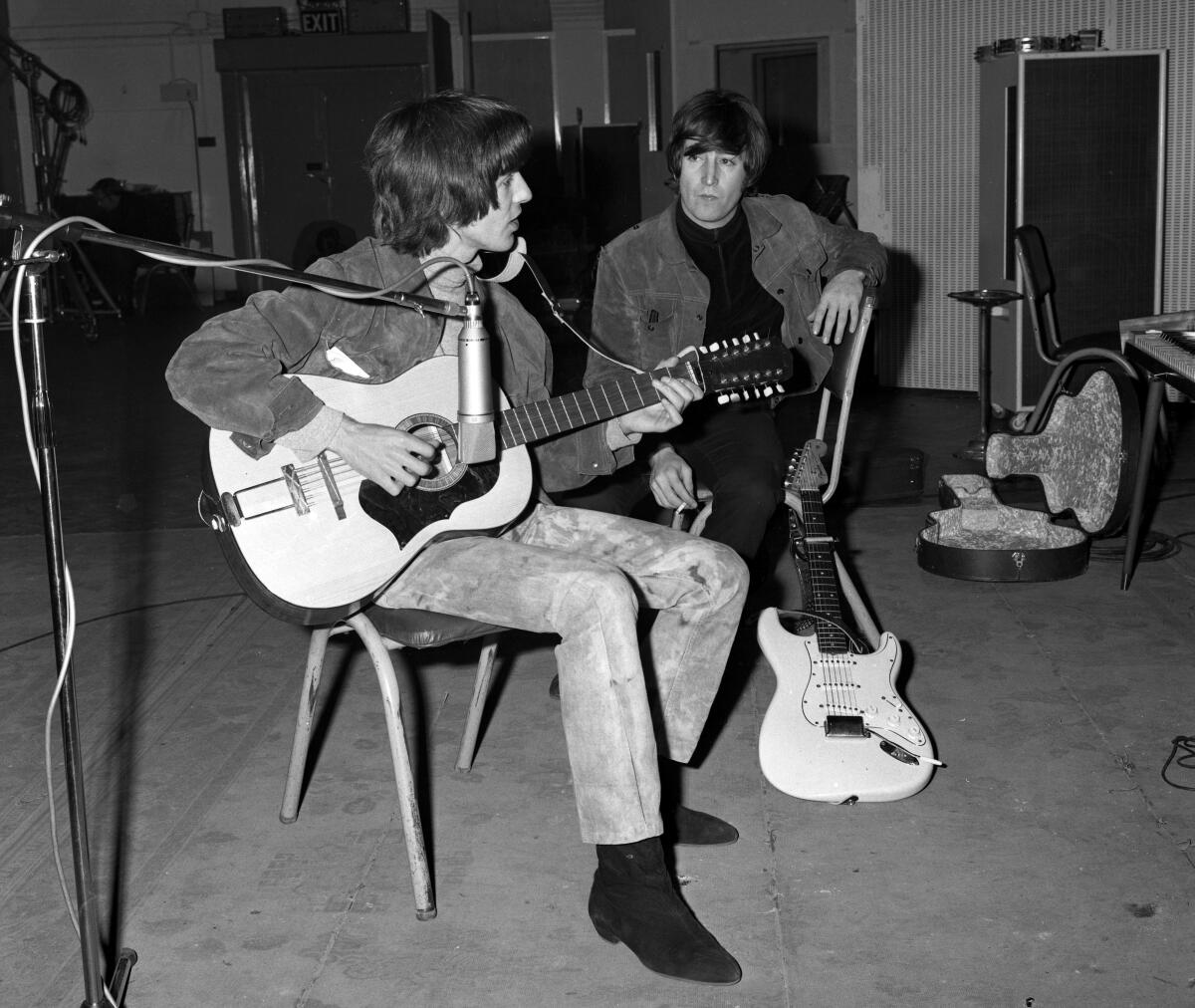 A young man with a shaggy hairstyle sitting and playing a guitar while another man watches with a guitar leaning on his chair