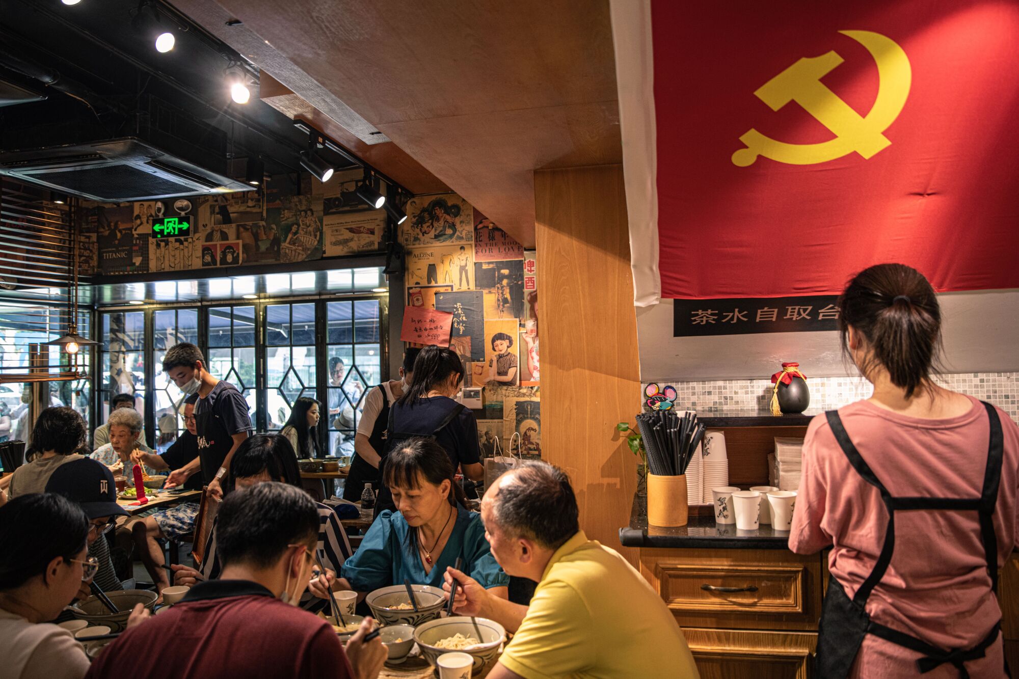 A red flag with a yellow hammer and sickle symbol hangs near people eating bowls of noodles at a table