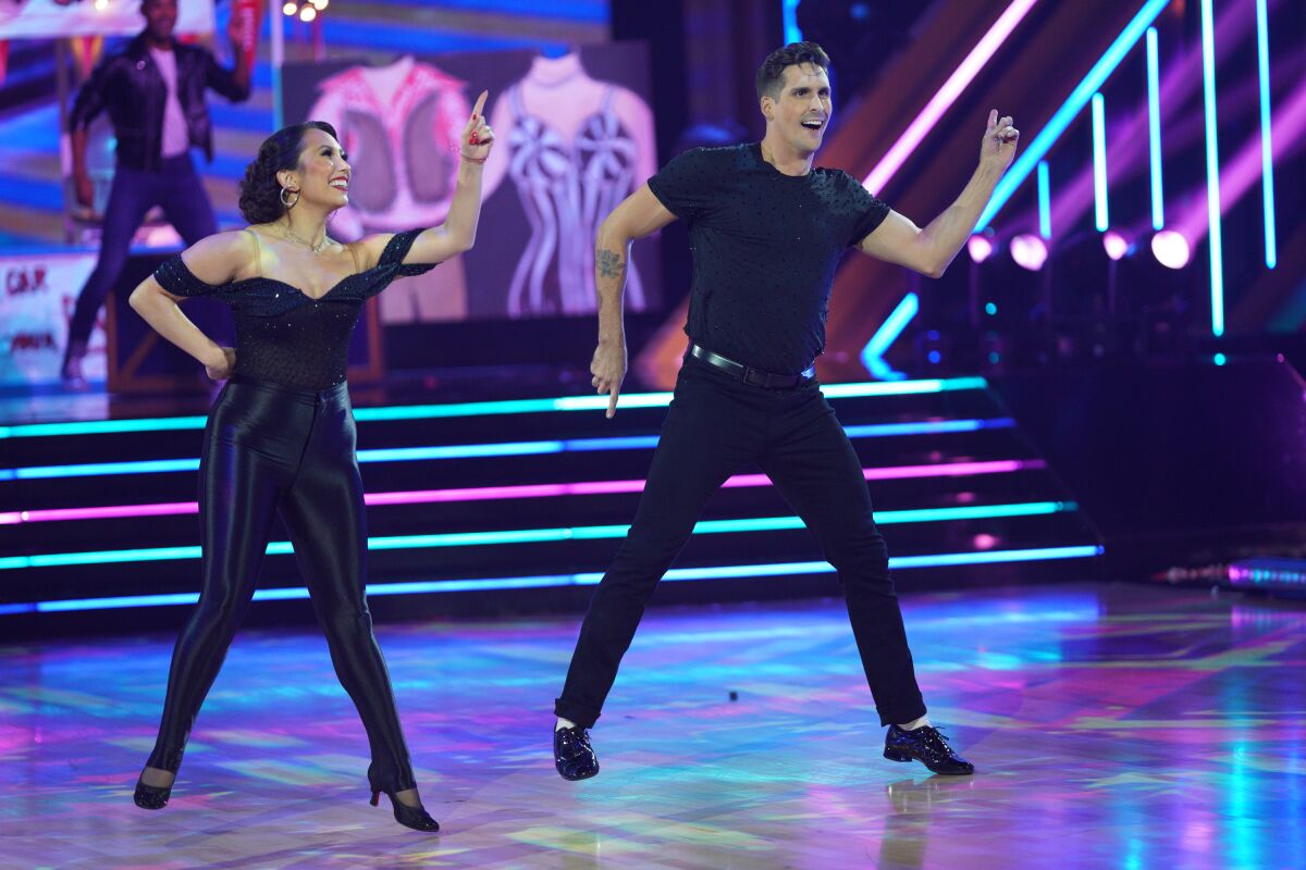 A man and woman in all black outfits inspired by "Grease" dance under colorful lights.