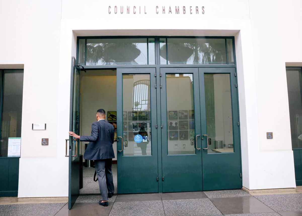 Wearing a suit, Triston Ezidore walks through the front door of a building with a sign saying "council chambers."