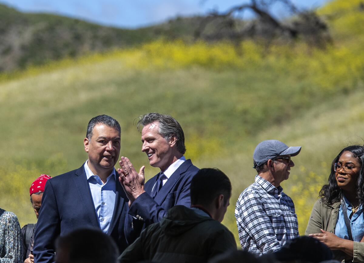 Two men in suit jackets stand among a small group on a grassy hillside.