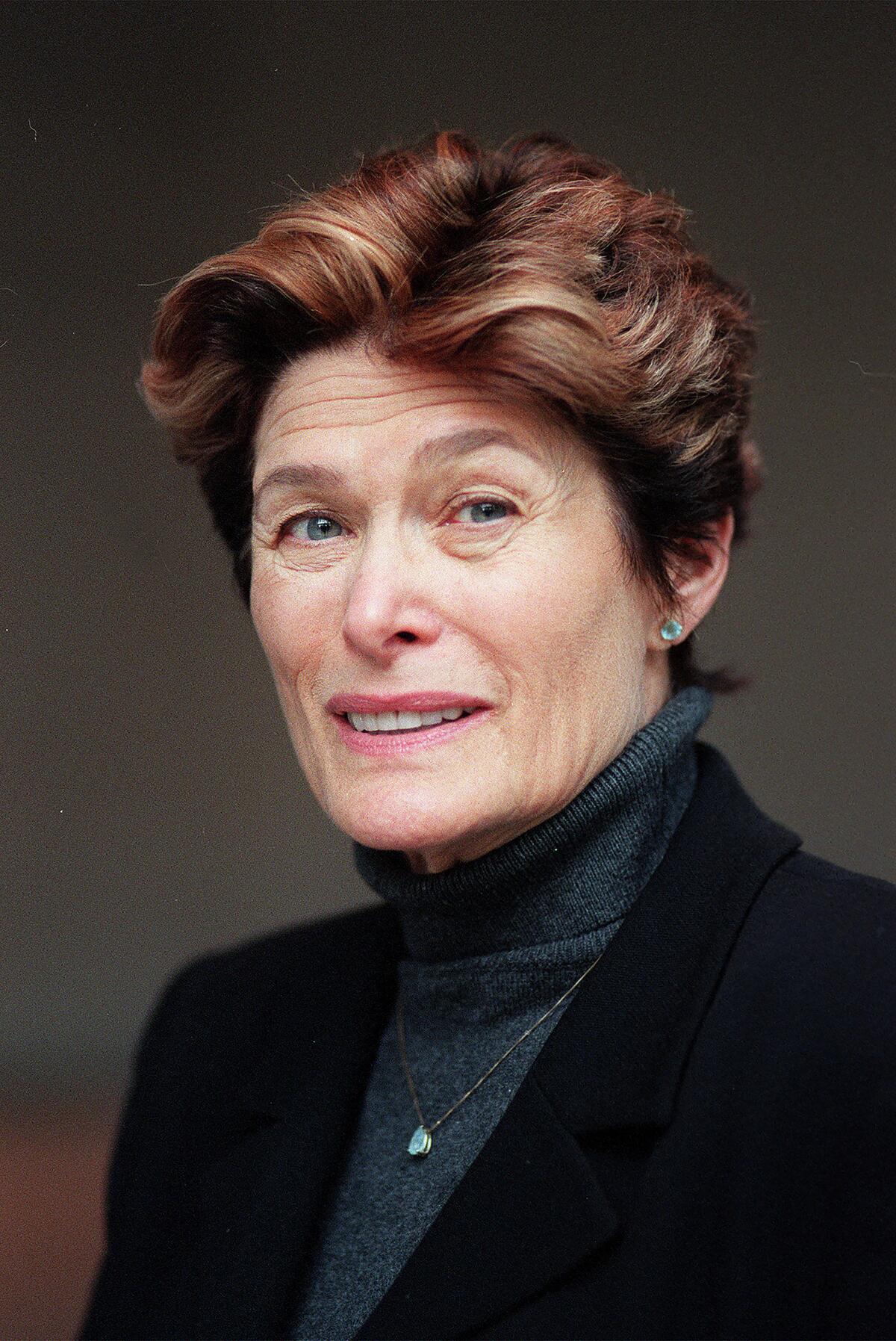 A woman with short brown hair, wearing a dark blazer over a turtleneck, smiles at the camera.