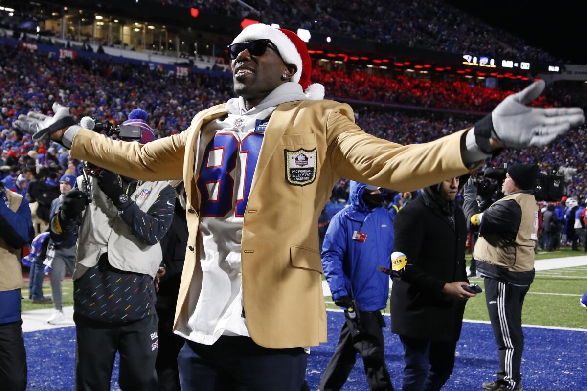 A man in a football jersey, sport coat and Santa hat spreads his arms standing in front of a small group on a football field