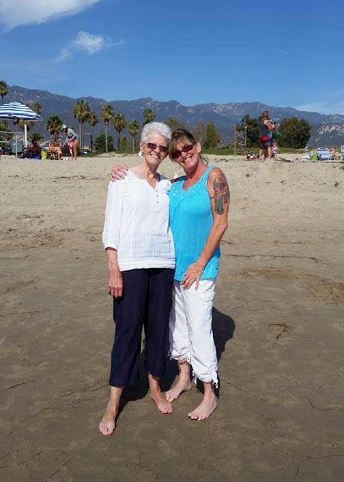 Two women press their heads together and wrap arms around each other while posing for the camera standing on a beach.