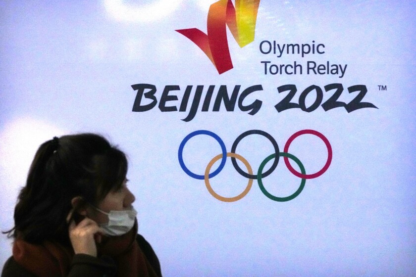A woman wearing a face mask to protect against COVID-19 walks past the logo for the Beijing 2022 Winter Olympic Torch Relay during an event at the Beijing University of Posts and Communications in Beijing, Thursday, Dec. 9, 2021. Already roiled by the pandemic and a partial diplomatic boycott, the Beijing Winter Olympics face another challenge in finding airplane seats for all the athletes and officials. (AP Photo/Mark Schiefelbein)
