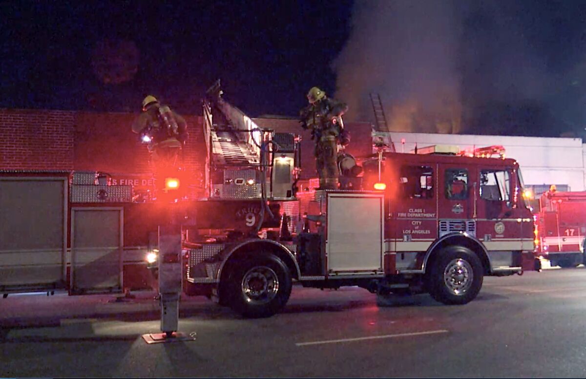 In the early morning, fire trucks are parked outside a fire in Boyle Heights, with a ladder extended over a roof.
