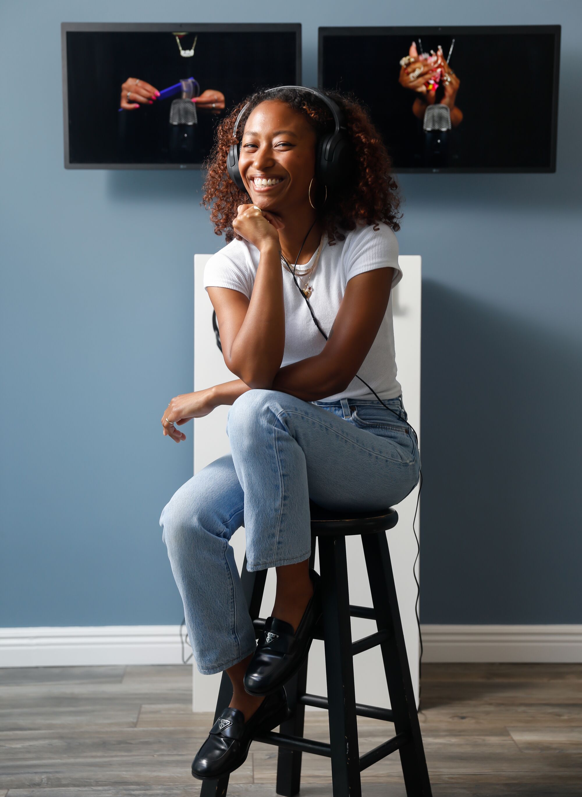 A Black woman wears headphones and sits on a stool smiling