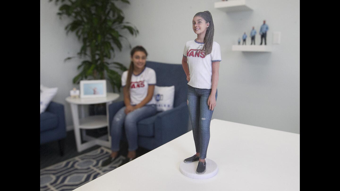 Sophia Warfield sits as her lifelike miniature of herself, a 3D "print" created with 3D photography and printing, stands on a table in front of her at 3d Likeables in Huntington Beach.