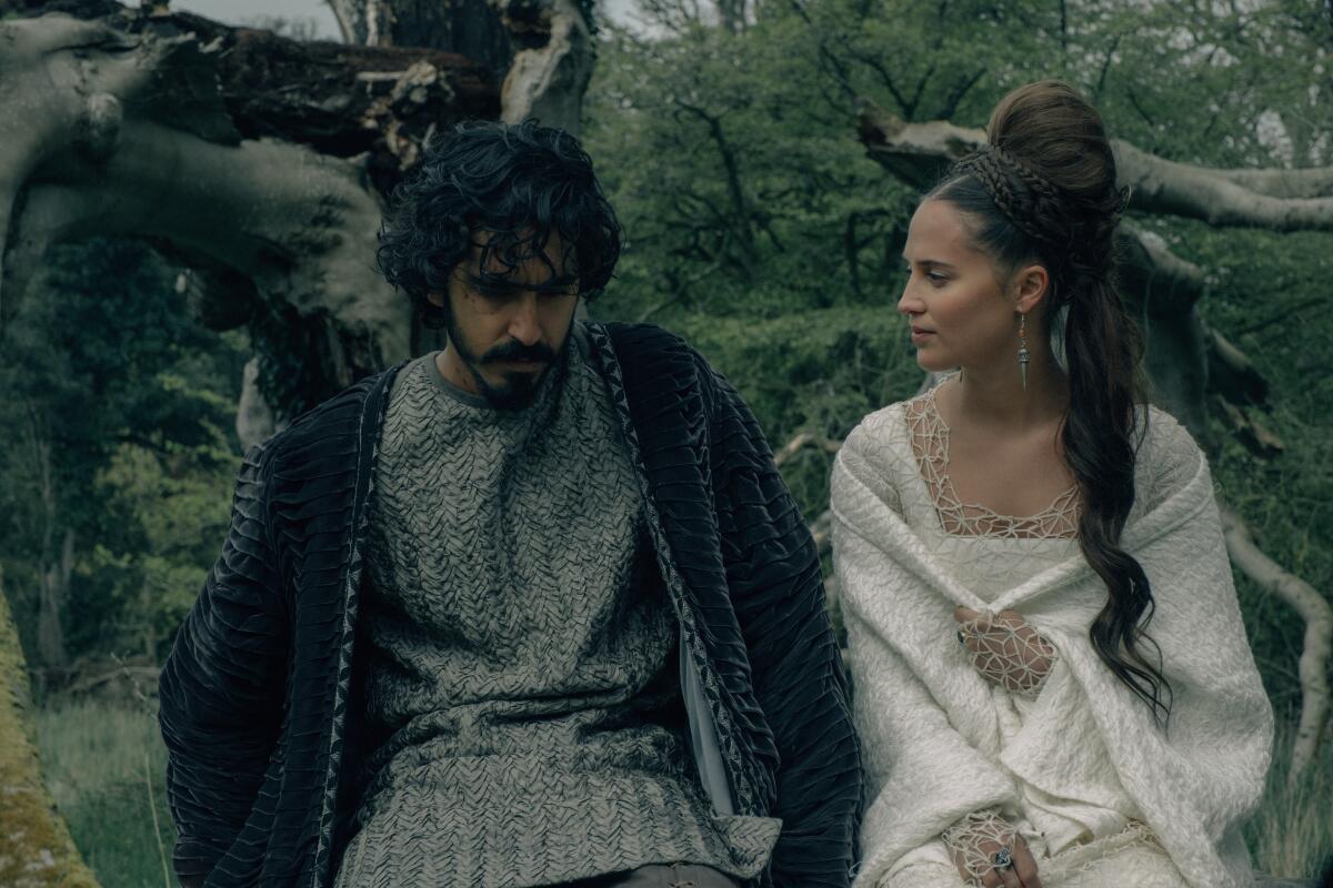 A man, left, and a woman in a white, 14th century-style dress sit in the woods. 