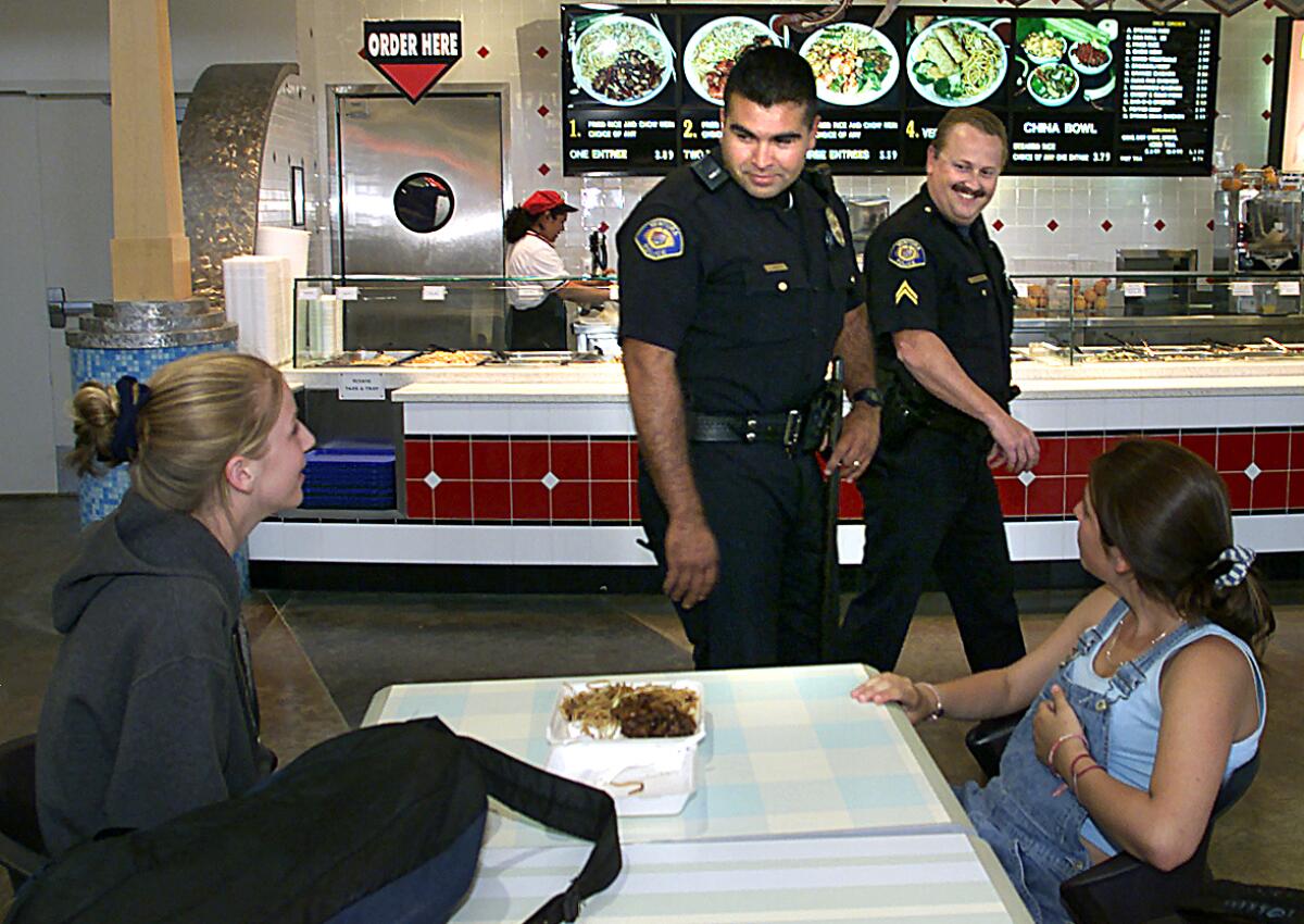 Two police officers in uniform walk next to a table at a mall food court and talk to two women sitting there