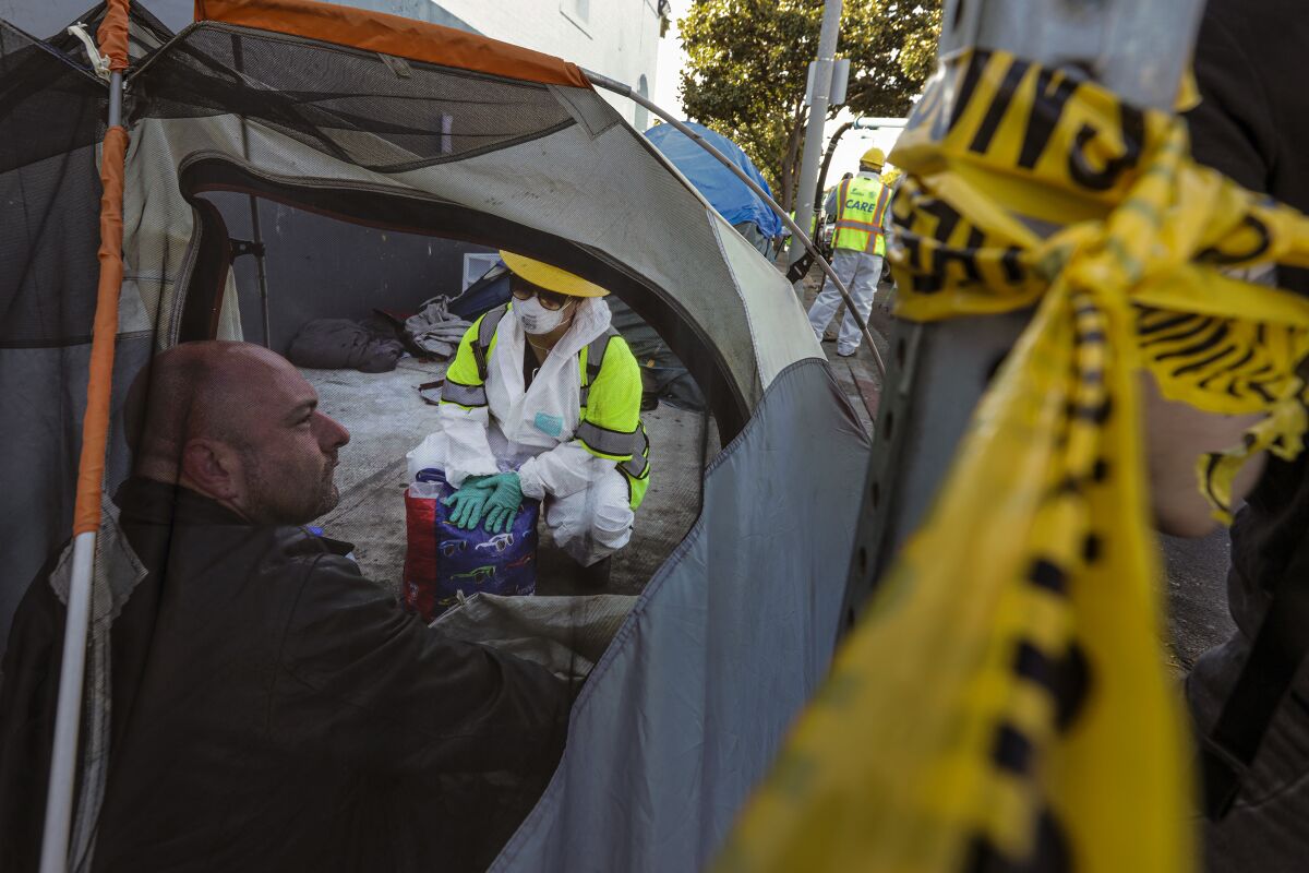 A city worker in full protective gear kneels to talk to a man inside a tent on a sidewalk in Hollywood