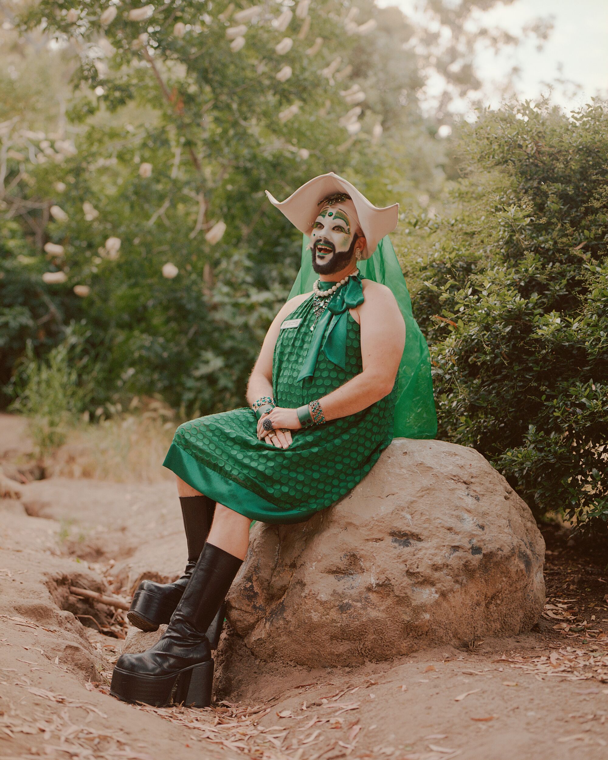 A drag nun in a green dress and boots sits on a boulder outdoors.