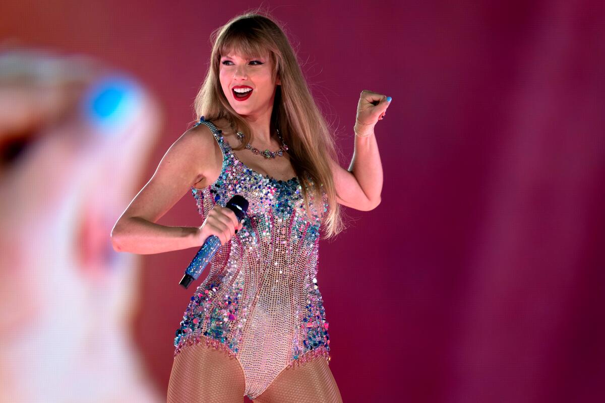 Taylor Swift stands on stage in a glittery leotard holding a mic in her right hand and making a fist with her left