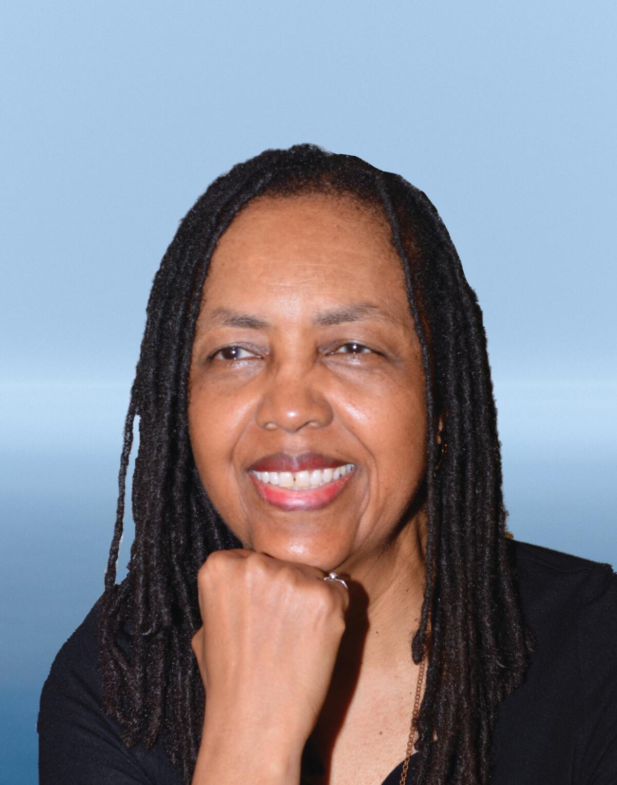 A woman with dreadlocks in a black long-sleeved shirt sits in front of a generic blue background for a portrait.