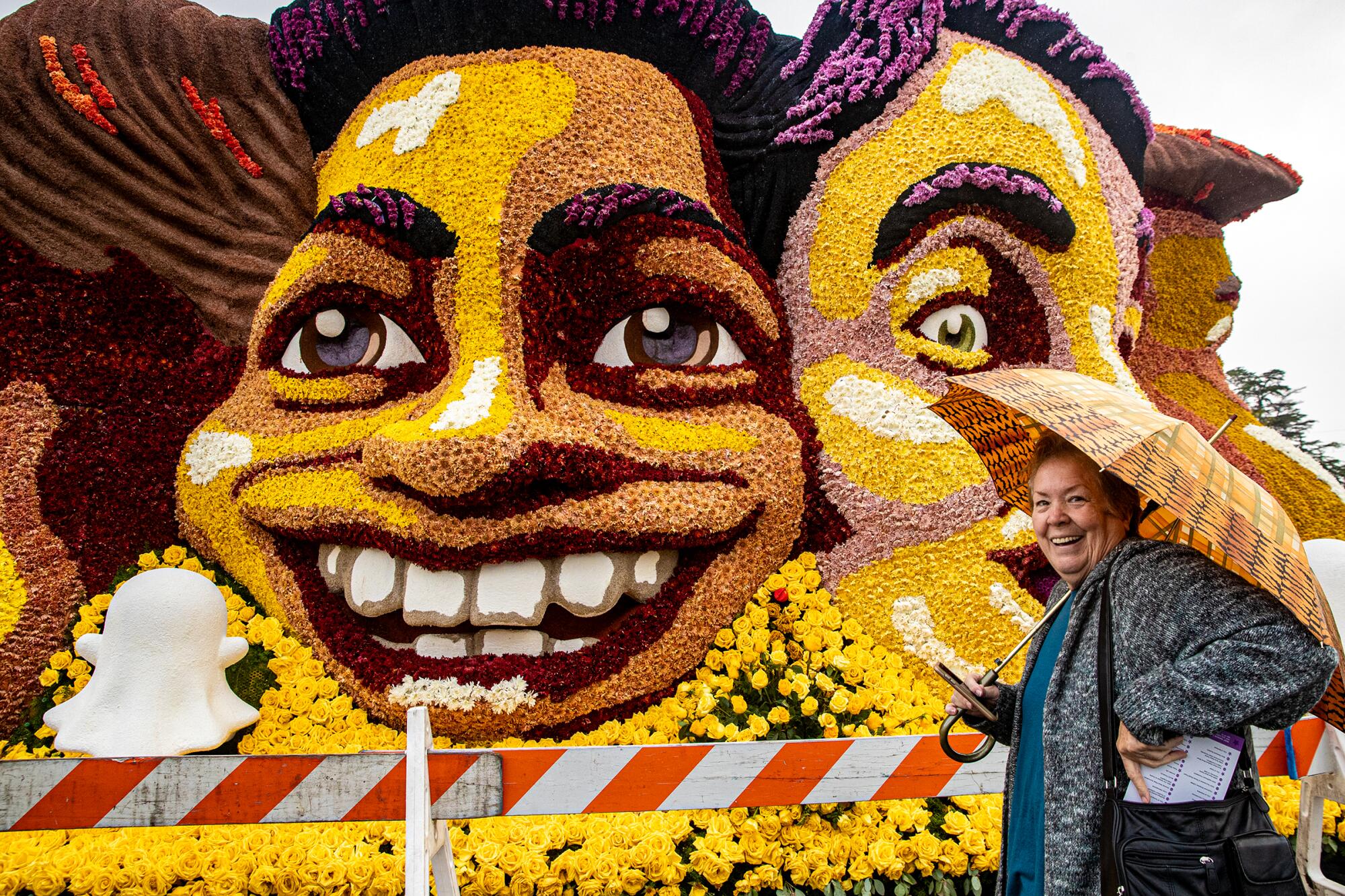 A woman stands and smiles with an umbrella next to a float with two faces 