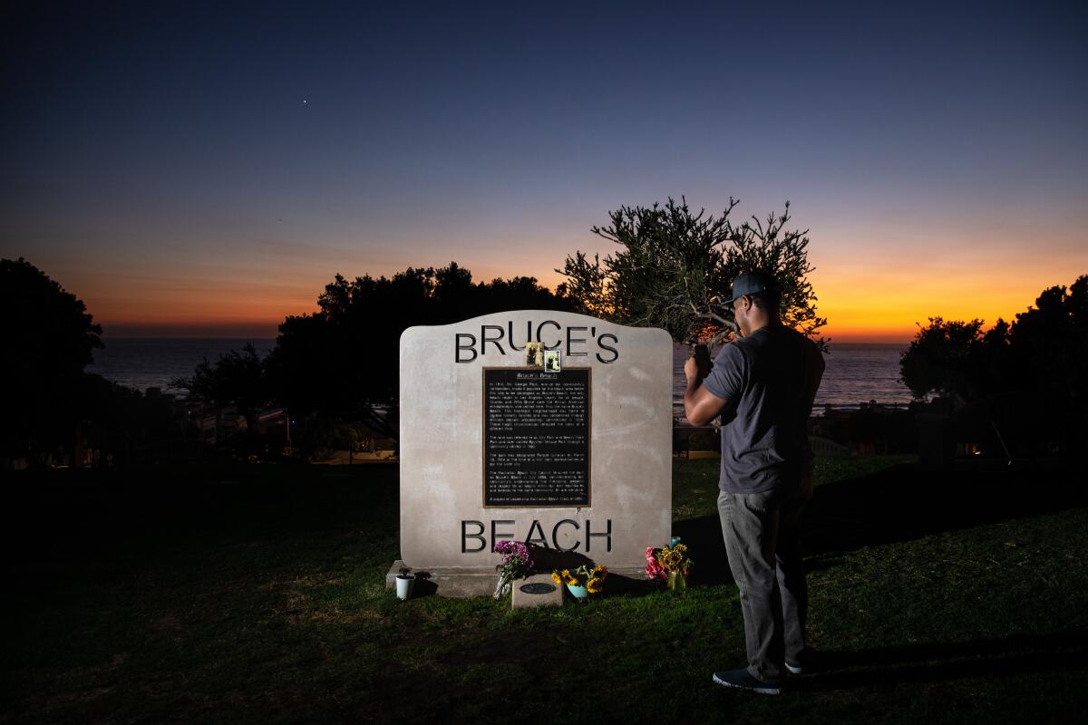 A man takes a picture of a plaque. In the background is the ocean at sunset.