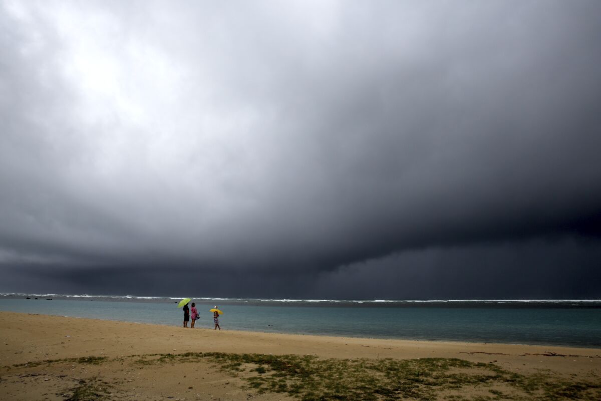 People hold umbrellas as it begins to rain on an otherwise empty beach in Honolulu on Monday, Dec. 6, 2021. A strong storm packing high winds and extremely heavy rain flooded roads and downed power lines and tree branches across Hawaii, with officials warning Monday of potentially worse conditions ahead. (AP Photo/Caleb Jones)