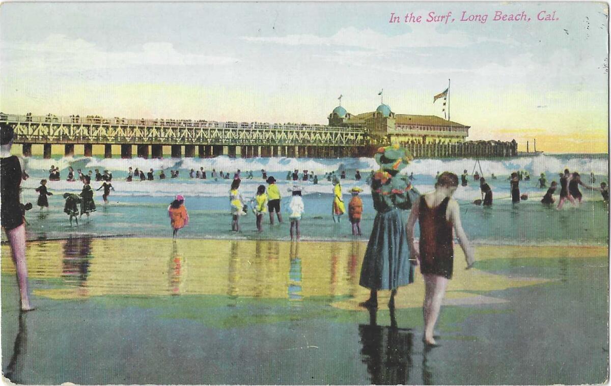 People play in the surf near a pier. Prominent in the frame is a woman in an ankle-length blue dress and a hat