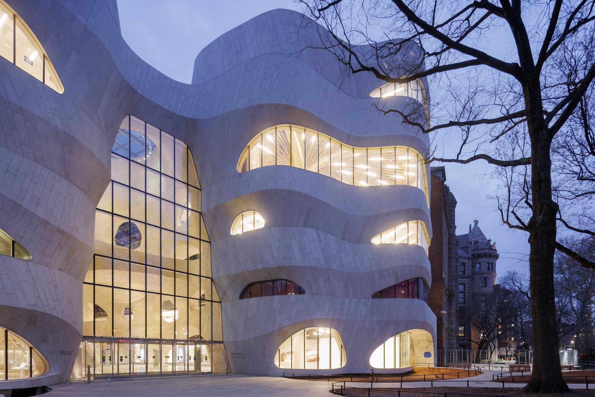 A building with a curving facade revealing irregularly shaped windows is illuminated from within at dusk