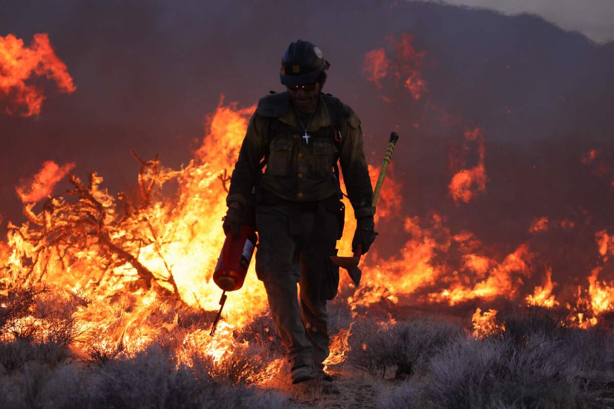 A firefighter holding an axe and a drip torch is silhouetted by the flames of a wildfire.