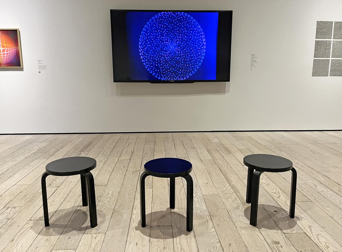 A view of a gallery shows three small black stools in front of a TV monitor displaying a small geometric pattern.