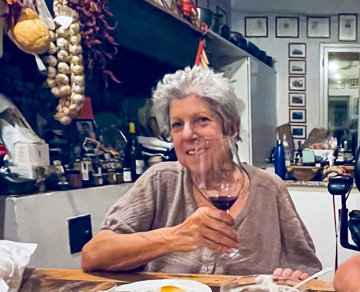 A woman sits at a kitchen table smiling as she lifts a glass of wine.