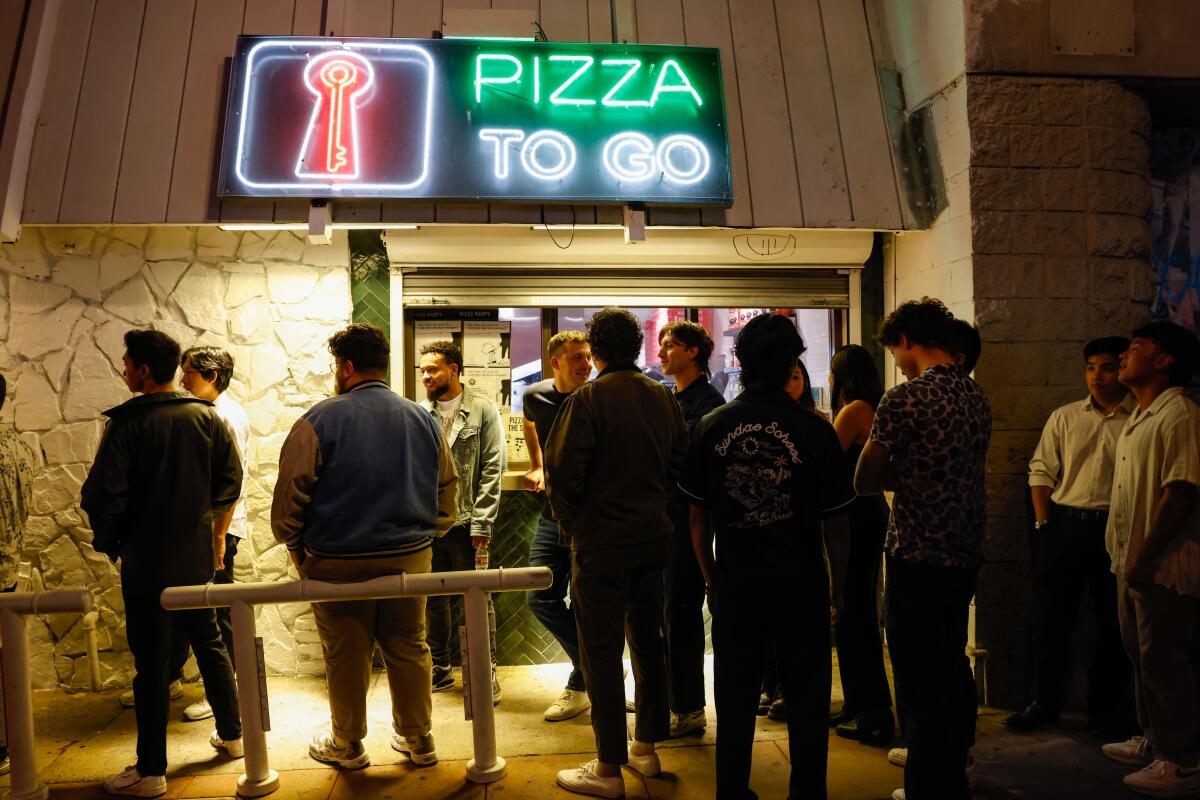 People stand outside a building with a neon sign on the wall that says Pizza to Go