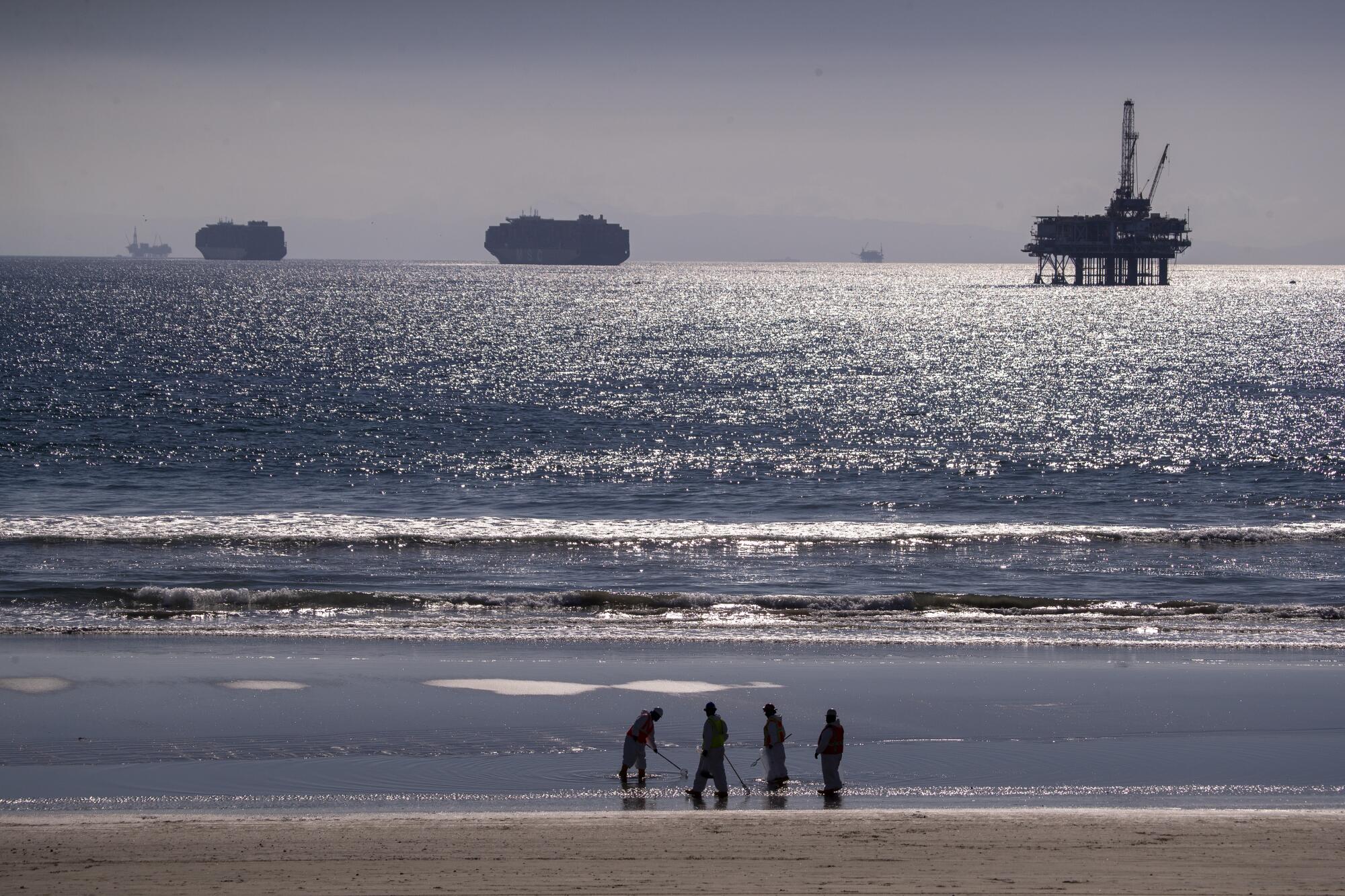 Container ships and an oil derrick line the horizon as oil spill cleanup crews work along the beach.