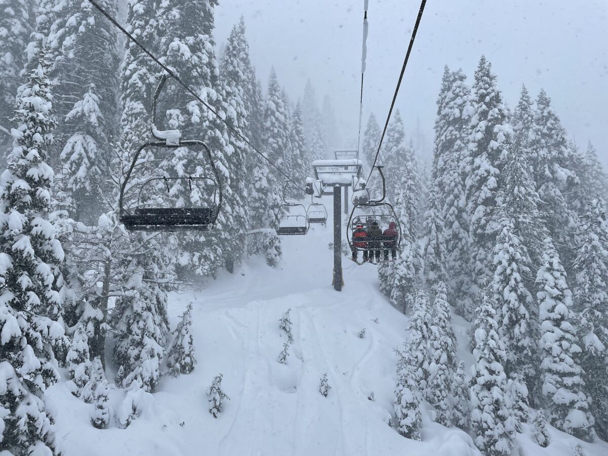 On a cloudy day, three people ride a ski lift over snow-covered evergreens.