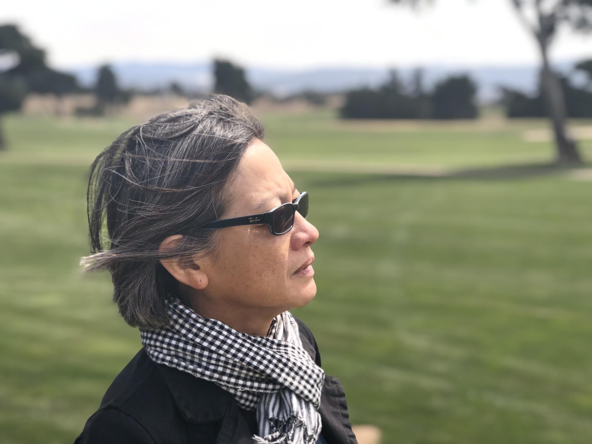 A woman wearing sunglasses and a checkered scarf pictured from the shoulders up in a grassy field, looking into the distance.