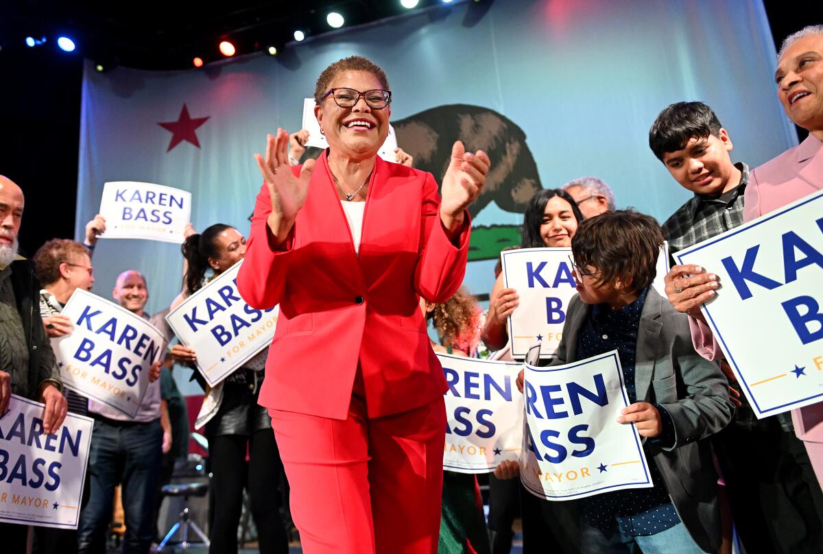 Karen Bass in a red suit claps while people behind her hold signs with her name