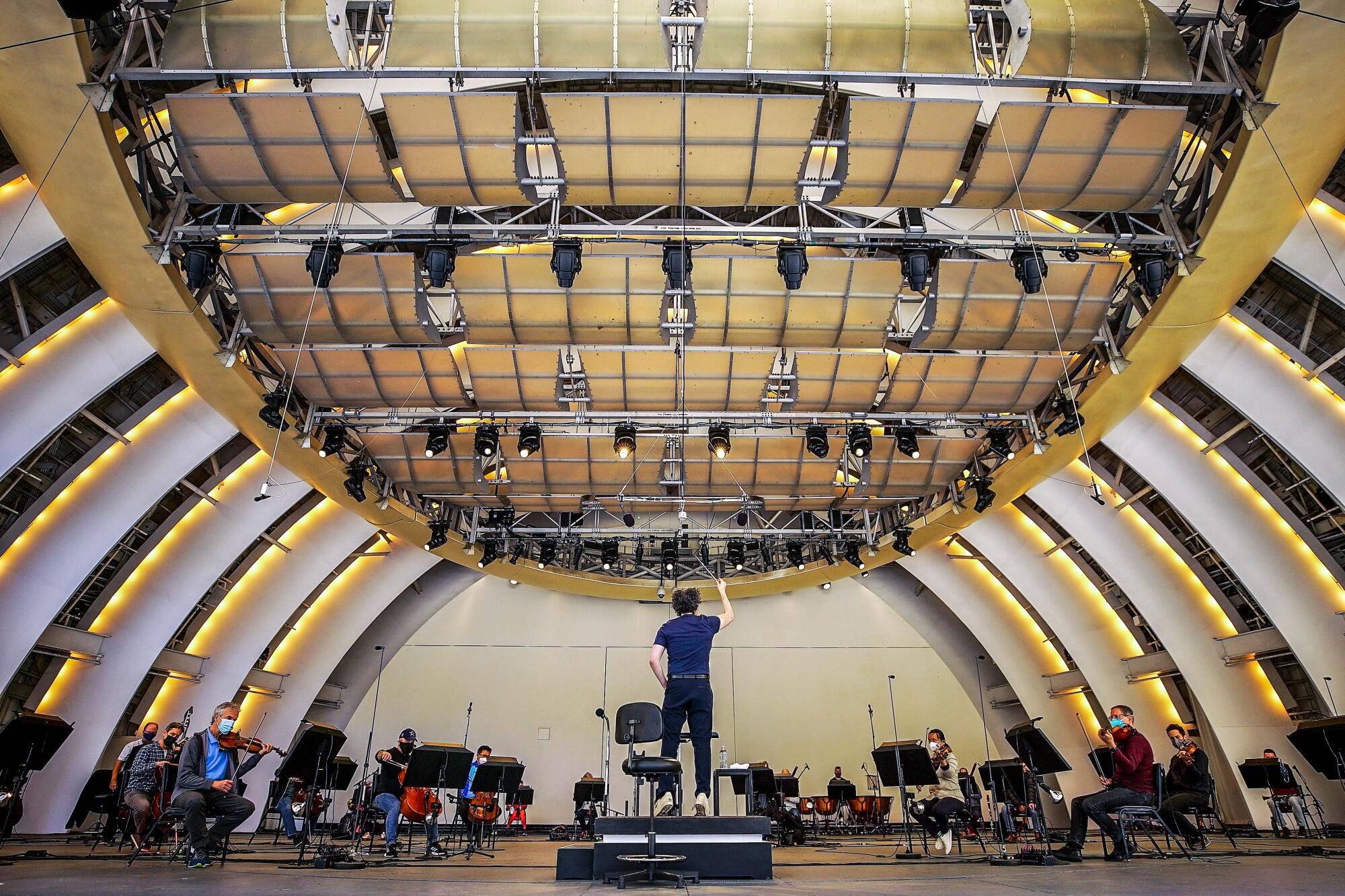 A wide view of a stage. A conductor in jeans bounces on his toes as he lifts a baton and masked musicians perform.