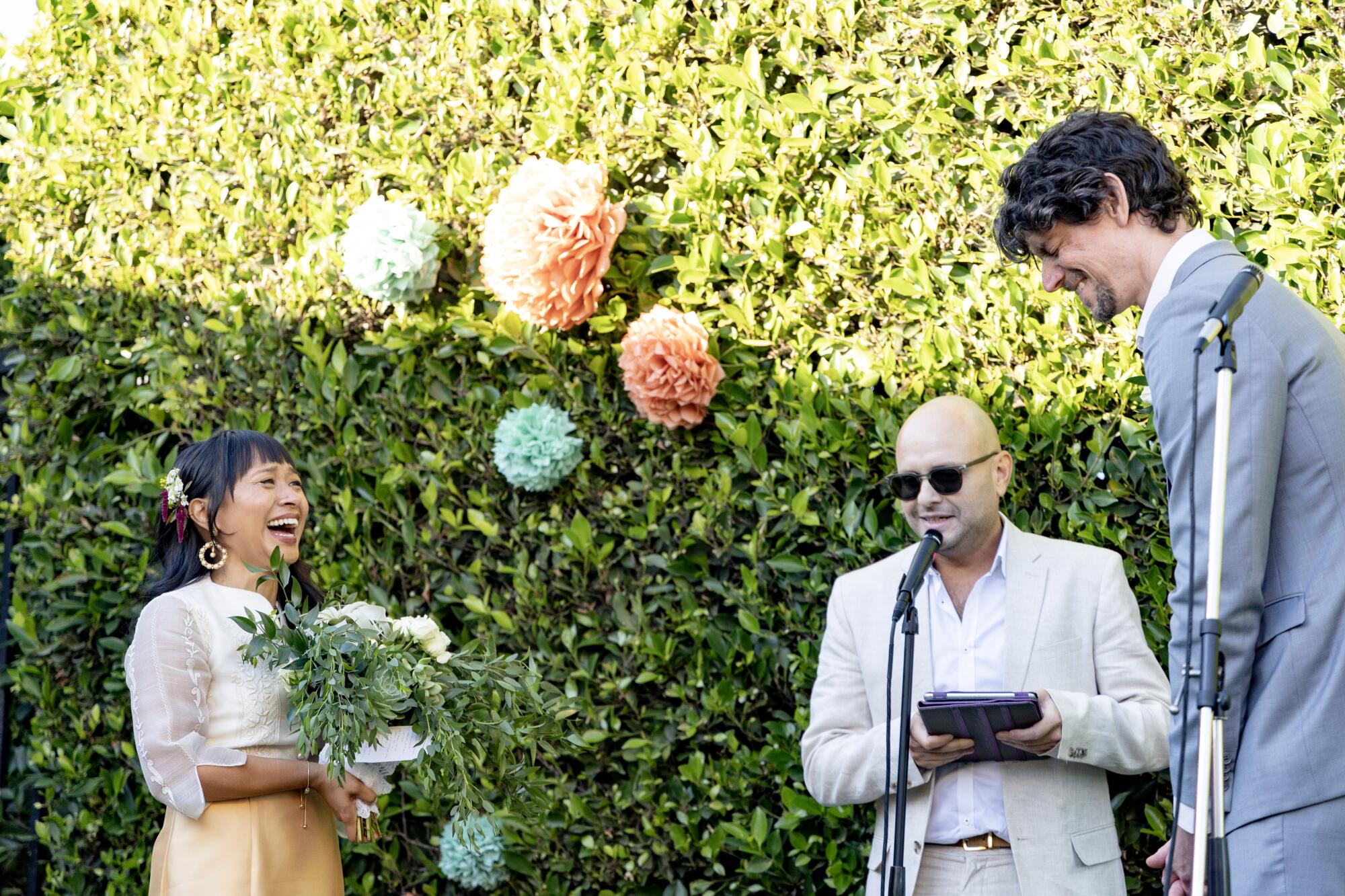 A laughing woman, a bald man in dark glasses and a tall man in a gray suit next to microphones in front of a hedge