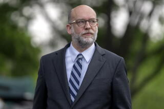A man with a graying beard, wearing glasses and a gray suit with a blue striped tie, looks to the right