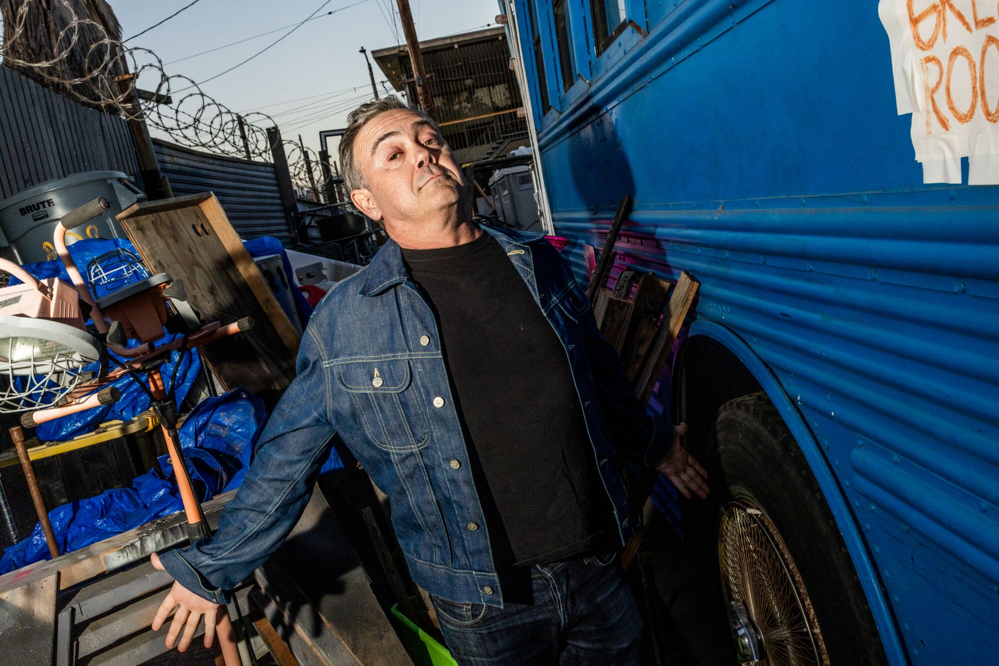 A man wearing a denim jacket poses outside a blue vehicle.
