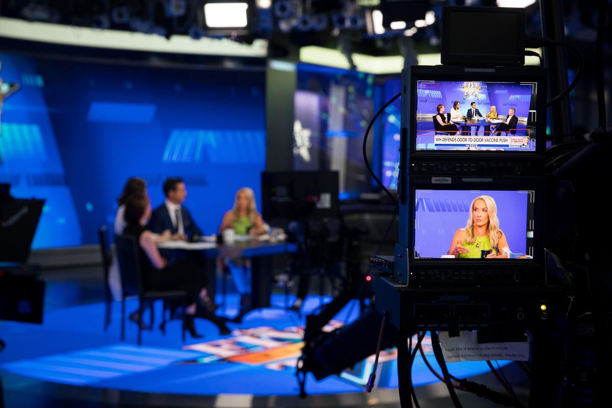 A woman is seen on a TV monitor as she and her show cohosts talk at a table in the background.
