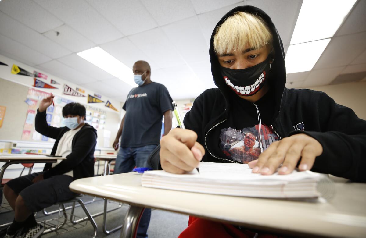 A student in a hoodie and a mask with grinning teeth works at a desk.