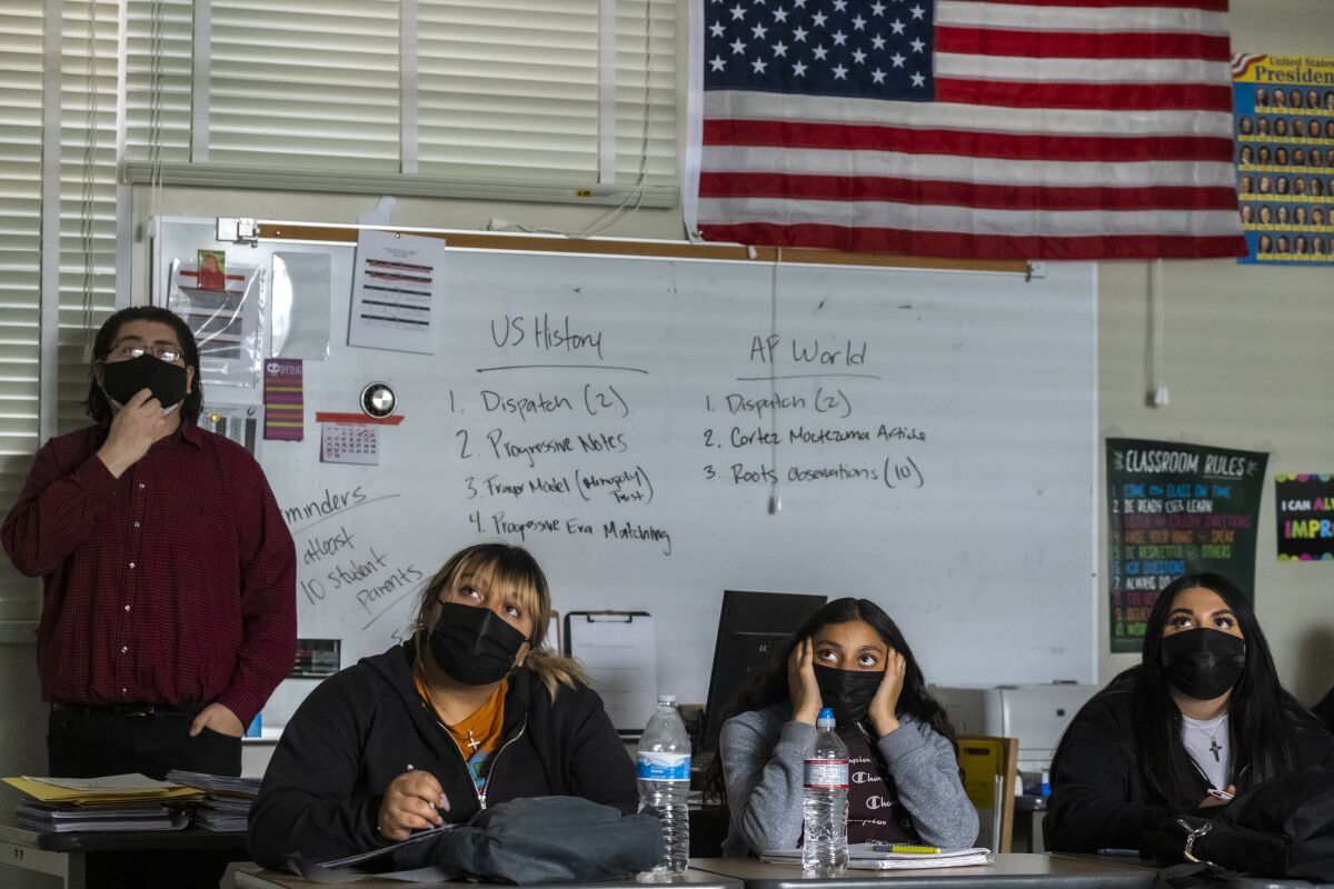 Four people wearing face masks in a classrom, one standing and three sitting.
