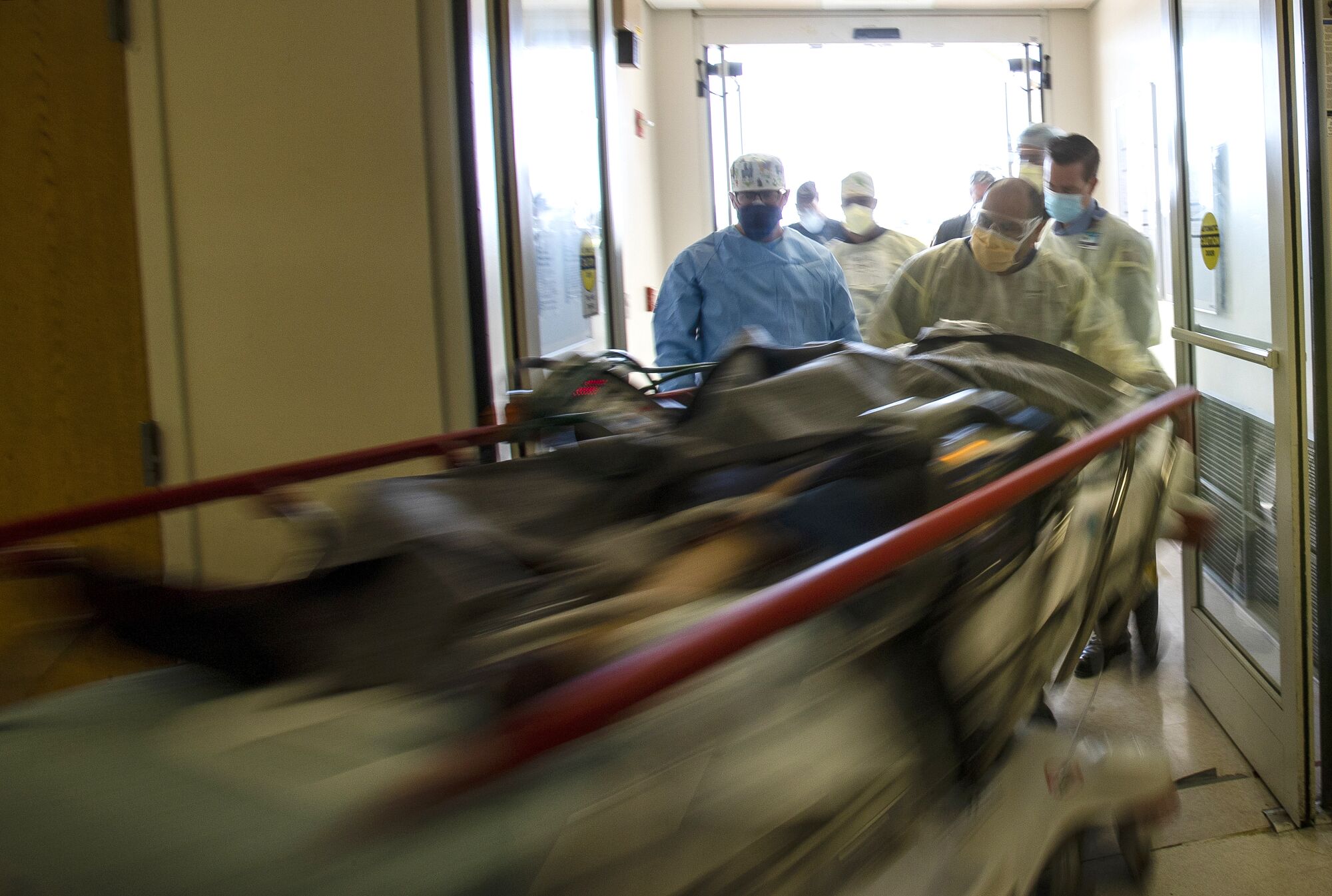 A hospital gurney streaks by, pushed by men wearing masks.