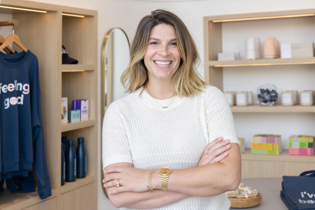 A woman in a store wearing a white shirt a gold watch crosses her arms and smiles