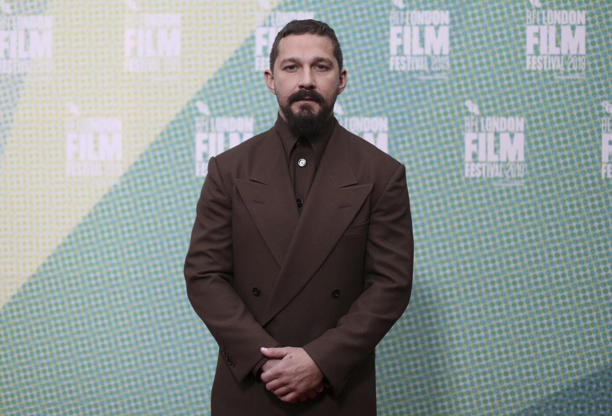 Actor Shia LaBeouf stands in a brown suit with hands clasped in front