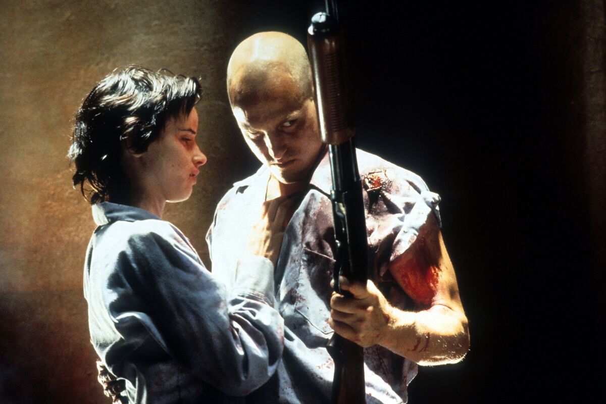 A woman is held by a bald man, who is holding a gun and wearing a short-sleeve shirt covered in blood.
