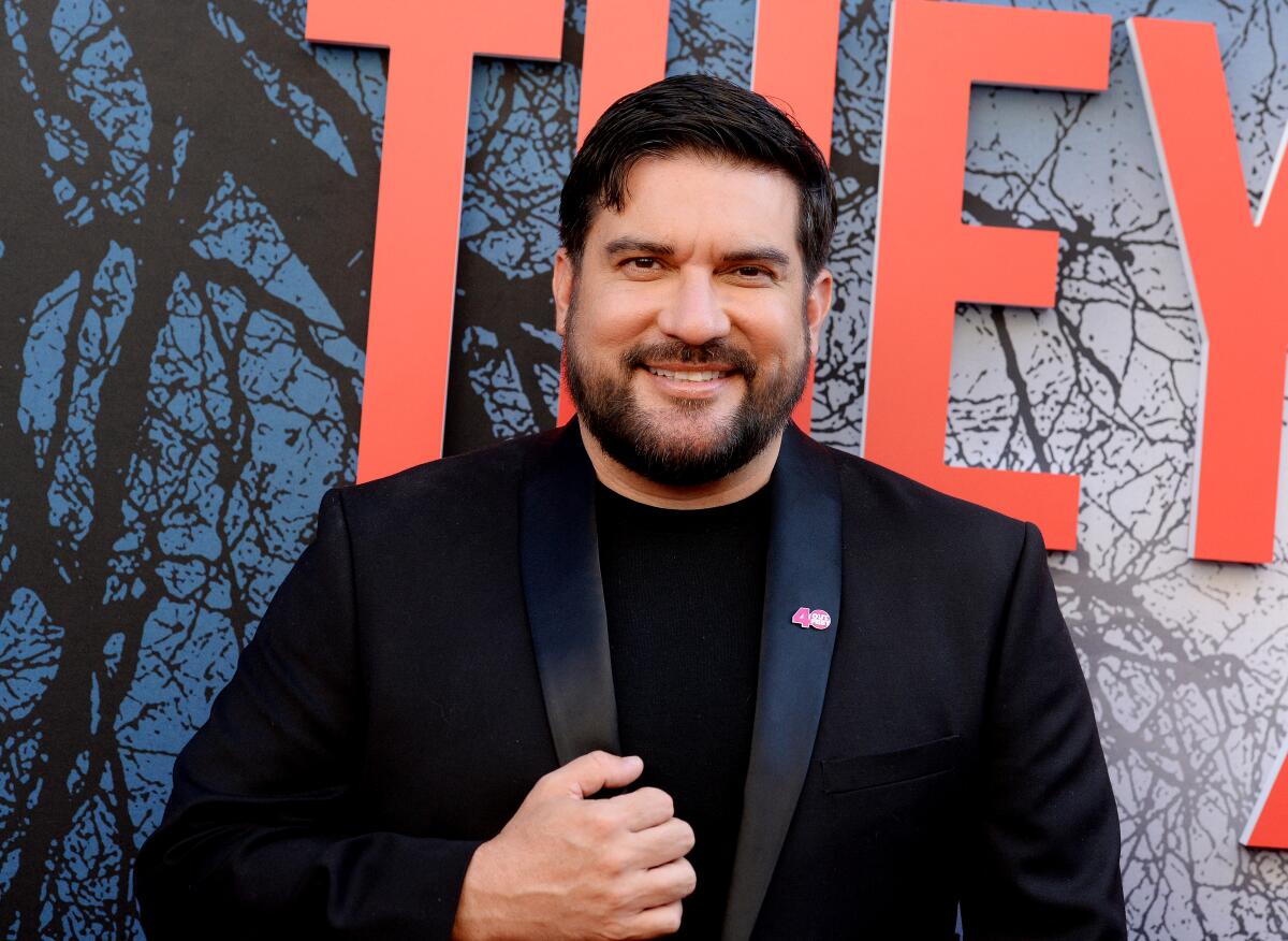 A smiling, bearded man in black suit jacket and T-shirt stands in front of a wall with the word "They" in red letters