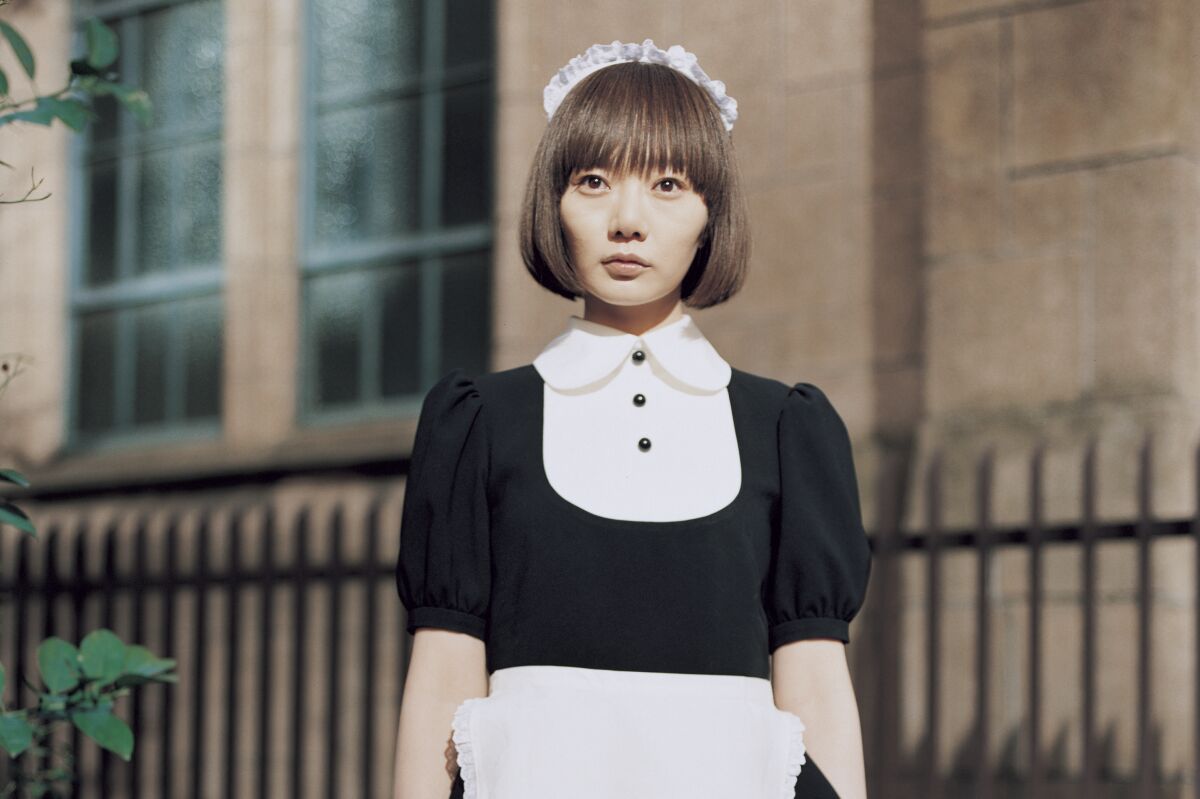 A woman dressed in a maid's uniform stands in front of a grand stone building.