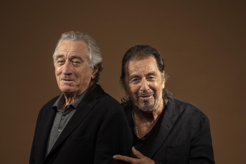 Robert De Niro wears a black blazer and stands next to Al Pacino who is dressed in black smiling, pointing to De Niro.