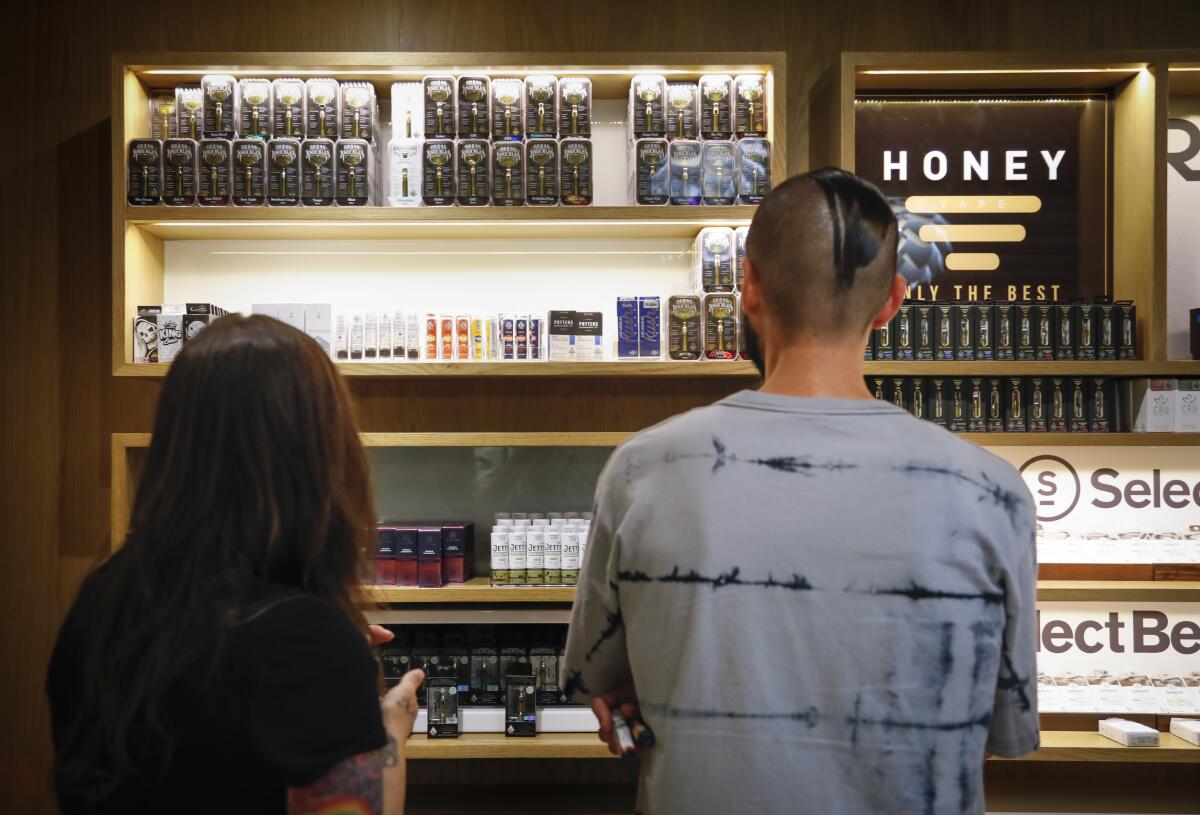 Two people are seen from behind looking at rows of cannabis products on shelves in a dispensary.