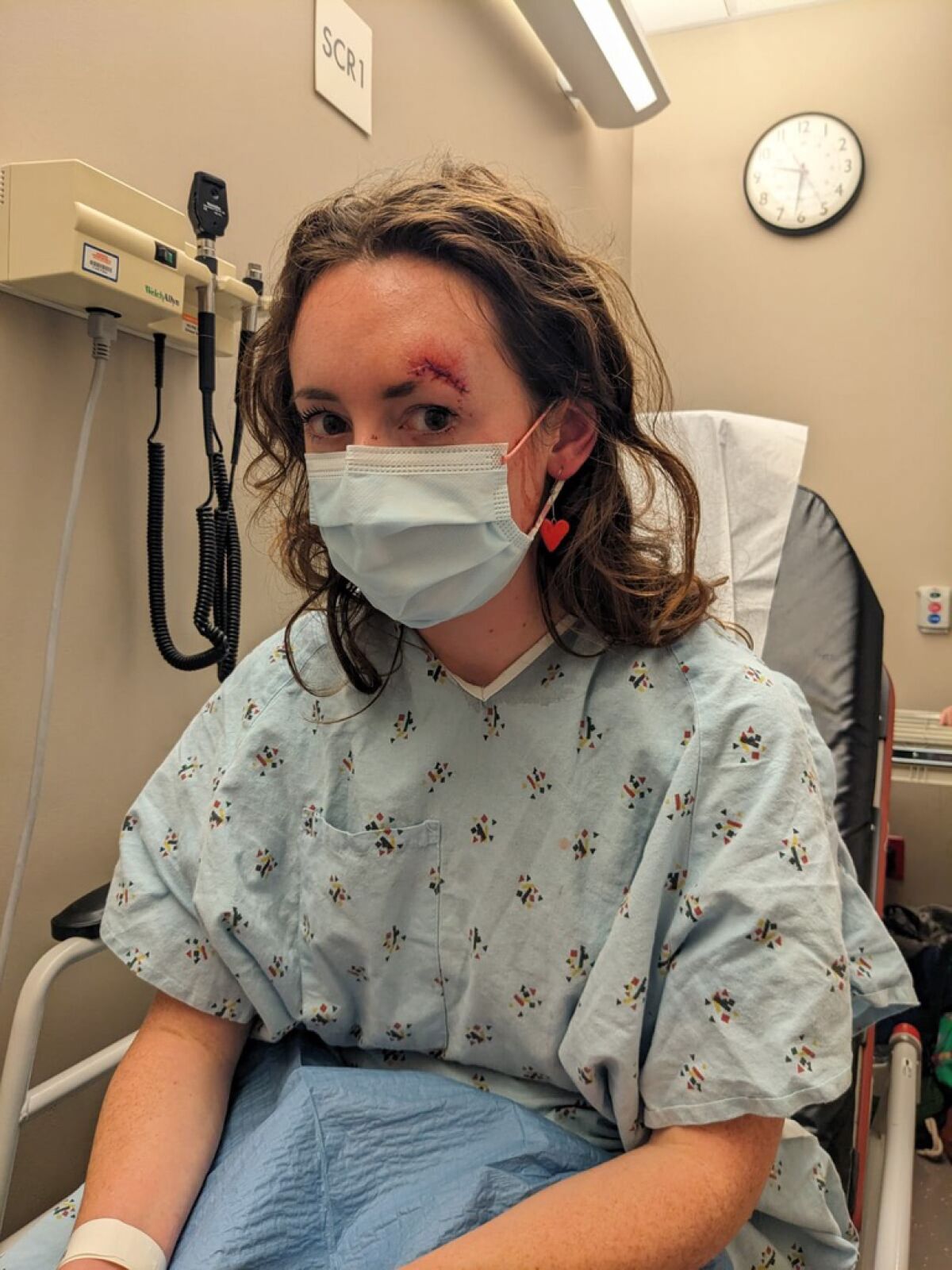 A woman in a hospital smock and face mask sitting in a medical room has stitches above one eyebrow.
