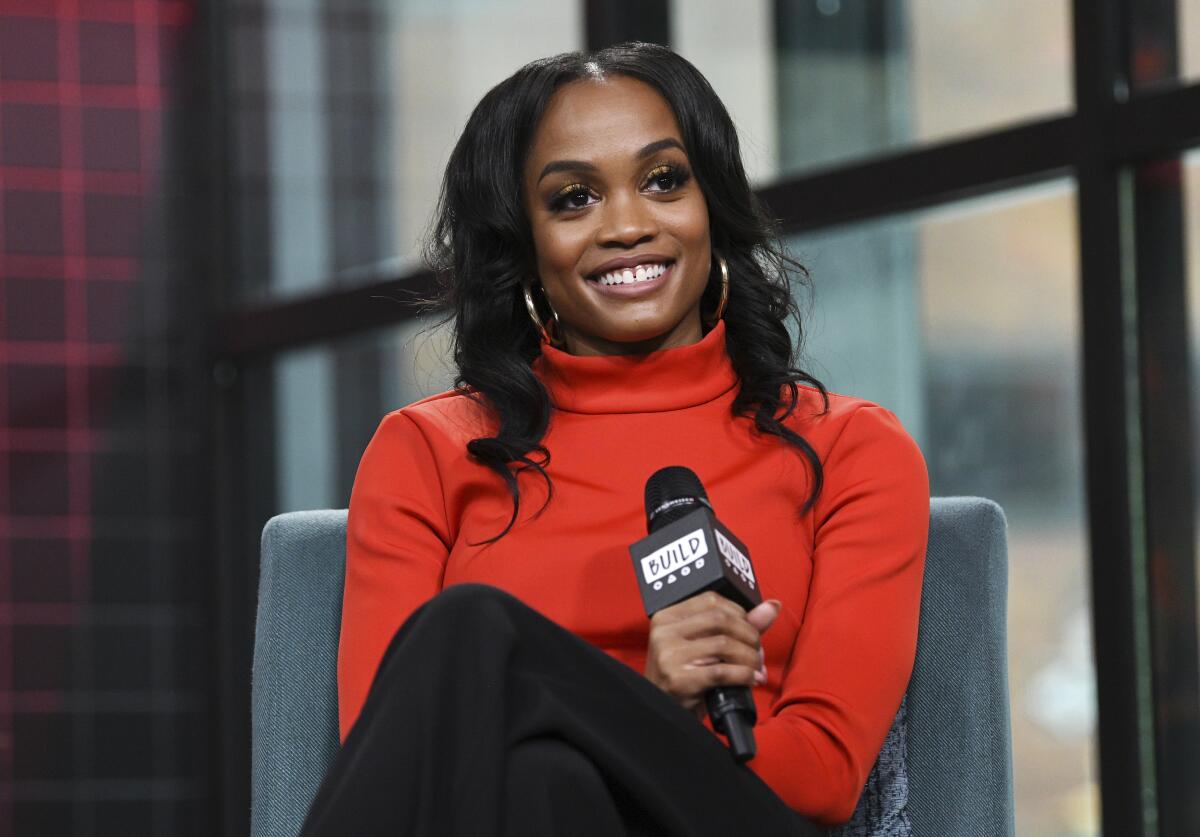 Rachel Lindsay in an orange turtleneck holding a microphone.