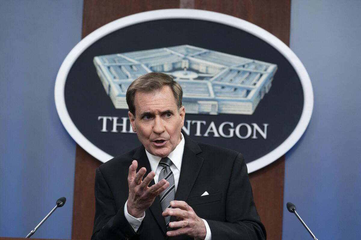 A man gestures with his hands while standing before microphones.